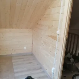 Poddasze wykończone boazerią z jasnego drewna, widoczne gniazdko elektryczne, listwy przypodłogowe i fragment podłogi z panelami, oraz widok na schody z drewnianą balustradą.