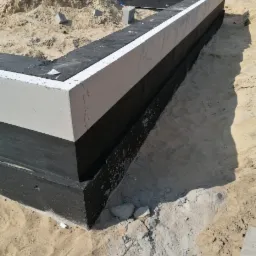 Wykop pod fundamenty z izolacją przeciwwilgociową i warstwą ocieplenia, widoczny piasek i elementy szalunku.