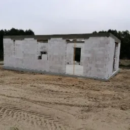 Surowy stan budynku z białych bloczków, widoczne fundamenty i ściany w trakcie wznoszenia, teren budowy z piaskiem i śladami opon.