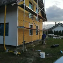 Dom w trakcie prac związanych z izolacją termiczną ścian zewnętrznych, widoczne rusztowania, materiał izolacyjny na elewacji, okna zabezpieczone folią, pracownik w oddali.