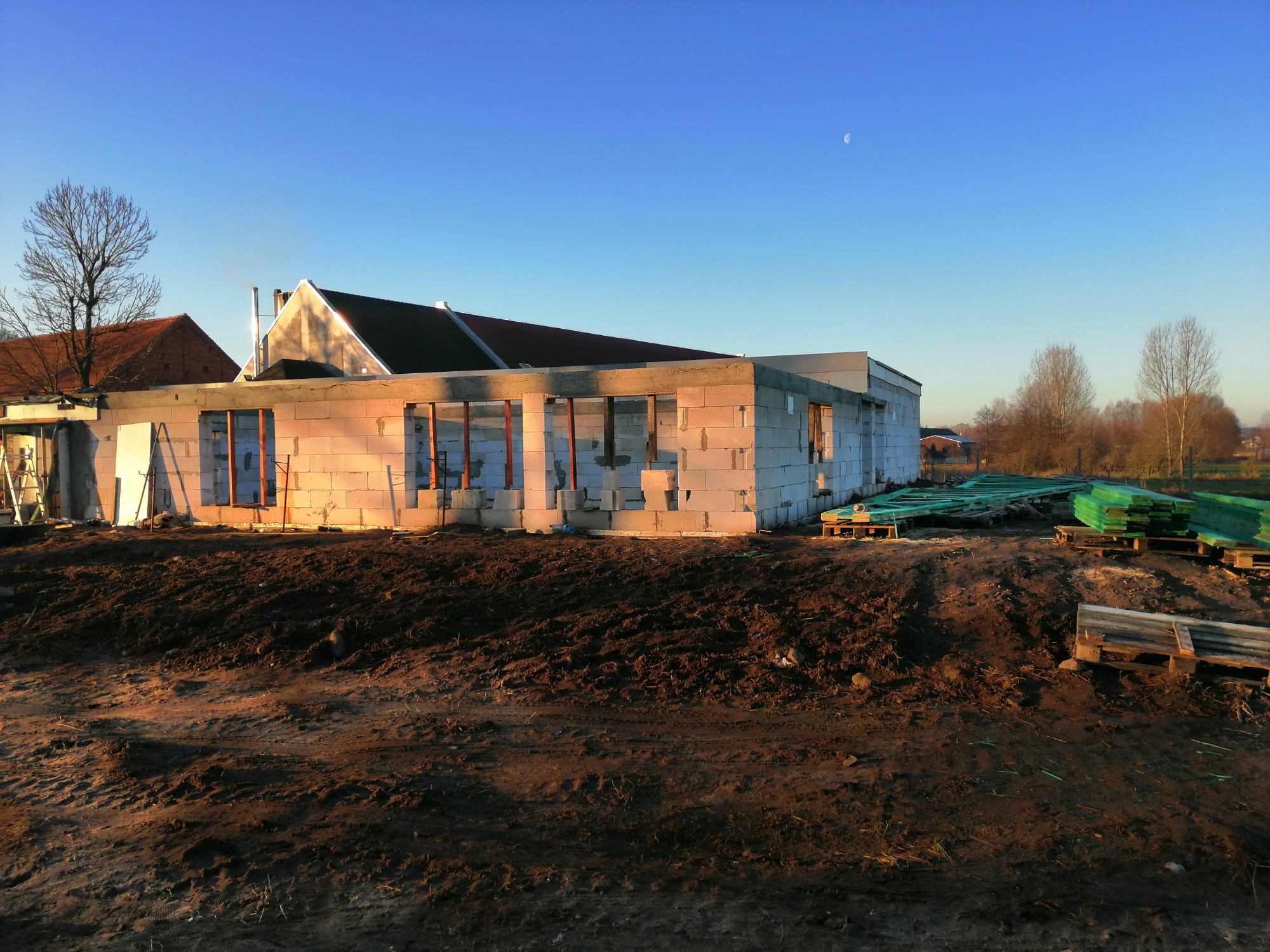 Surowy stan budowy domu z białych bloczków, widoczne otwory okienne i drzwiowe, palety z zielonym materiałem budowlanym na ziemi, w tle budynki i drzewa, błękitne niebo z małym księżycem.