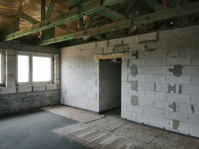 Wnętrze surowego domu w trakcie budowy: ściany z białych bloczków, drewniana konstrukcja dachu, okna z widokiem na zewnątrz, betonowa wylewka podłogi.