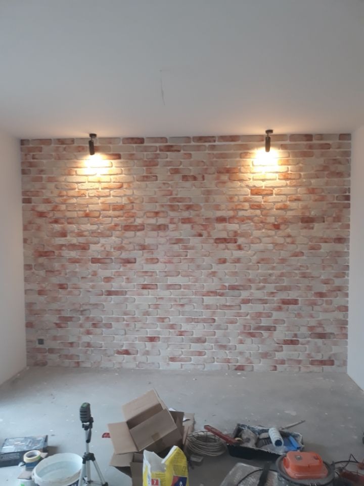 Ściana z cegieł dekoracyjnych, oświetlona dwoma punktowymi lampami, z widocznymi narzędziami i materiałami budowlanymi na podłodze w trakcie prac wykończeniowych.