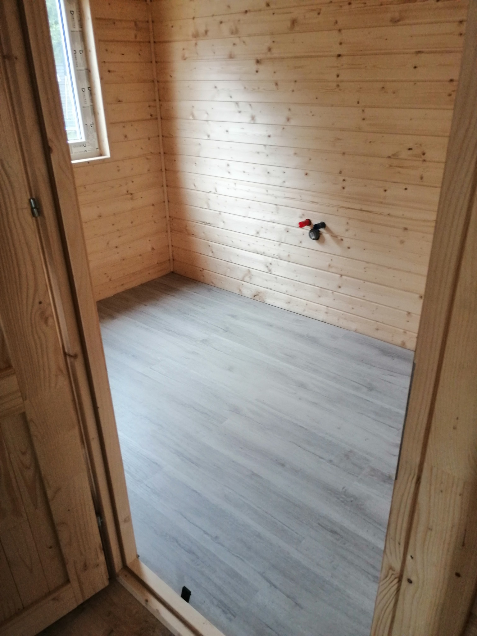 Wnętrze pomieszczenia wykończonego boazerią i panelami podłogowymi w odcieniach szarości, widoczne przyłącze wodne na ścianie i okno.