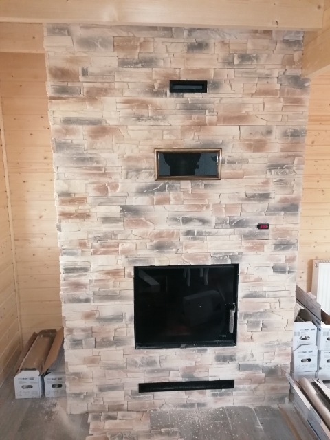 Ściana kominkowa wyłożona kamieniem dekoracyjnym w odcieniach beżu i szarości, z widocznymi elementami wentylacyjnymi i wkładem kominkowym, obudowana drewnianą konstrukcją.