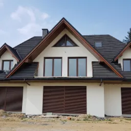 Nowoczesny dom jednorodzinny z białą elewacją, ciemną dachówką i brązowymi okiennicami w trakcie wykończenia, widoczne pozostałości po pracach budowlanych na podwórku.