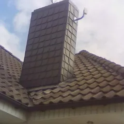 Dach pokryty brązową dachówką ceramiczną z obróbką komina w tym samym stylu, widoczny fragment orynnowania i wiatrowskaz na szczycie komina.