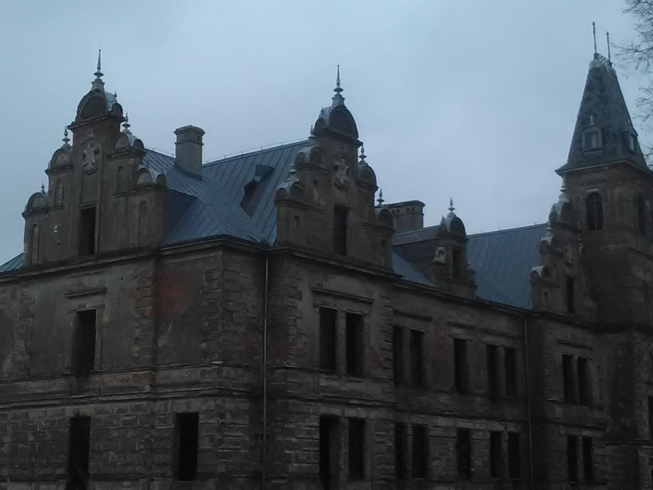 Zrujnowany, stary zamek z ciemną, nową blachą na dachu, widoczne kominy i wieżyczki, pochmurne niebo w tle.
