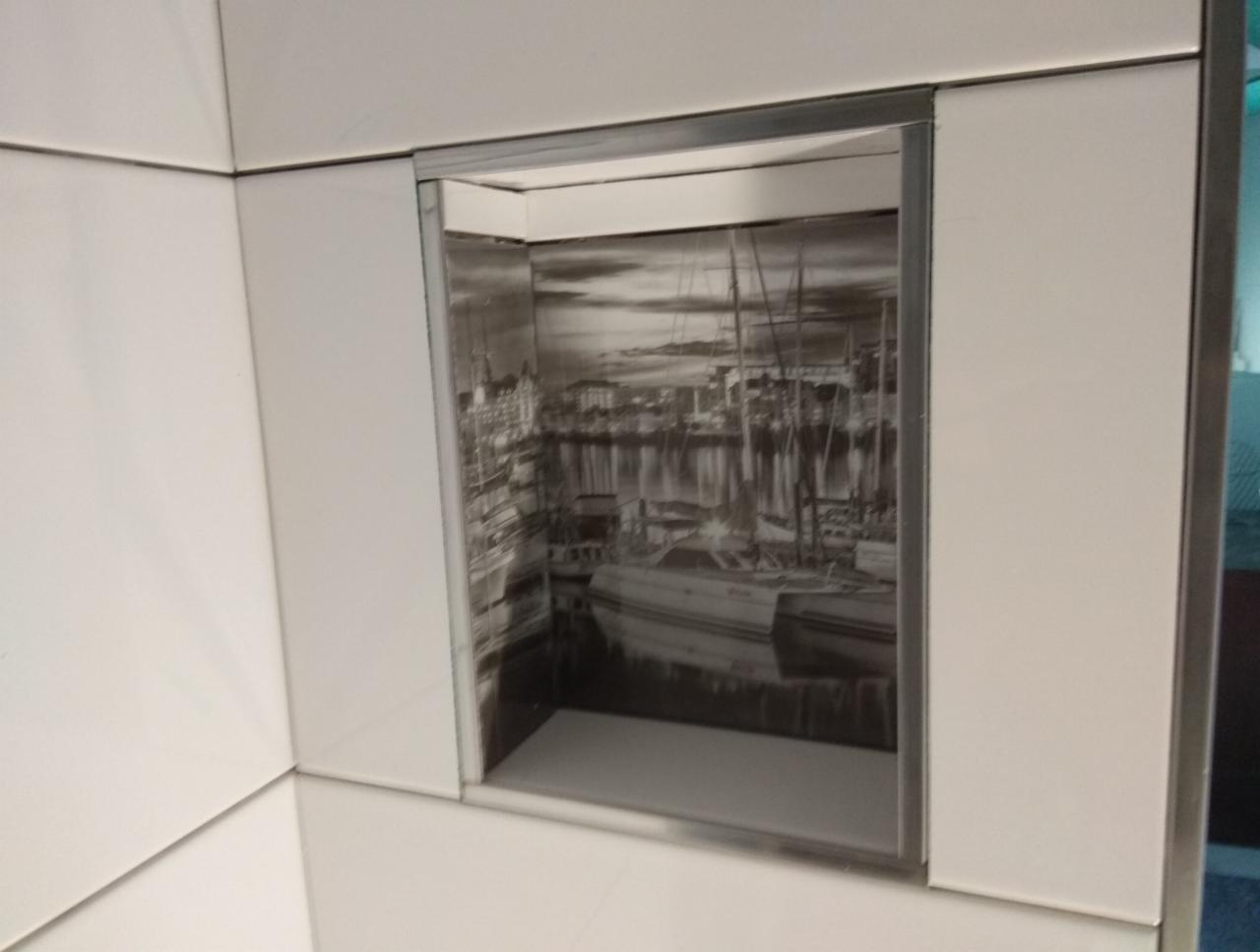 Białe, błyszczące płytki ceramiczne z wnęką, w której umieszczono czarno-białą grafikę przedstawiającą port z żaglówkami.