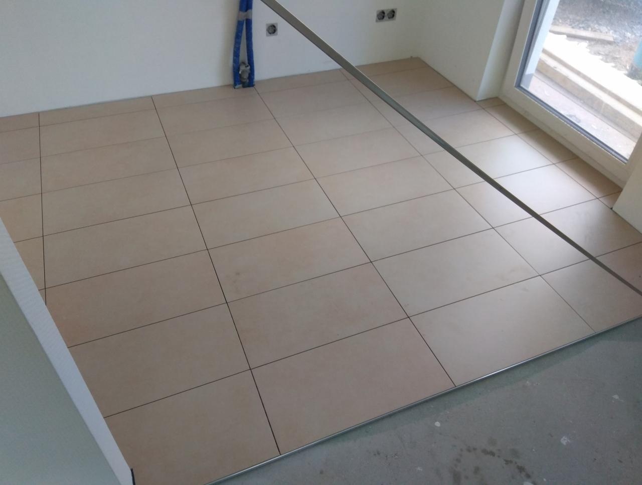 Ułożona podłoga z beżowych płytek ceramicznych z wąskimi fugami, widoczne rury instalacji wodnej przy ścianie oraz metalowy profil dylatacyjny przy oknie balkonowym.