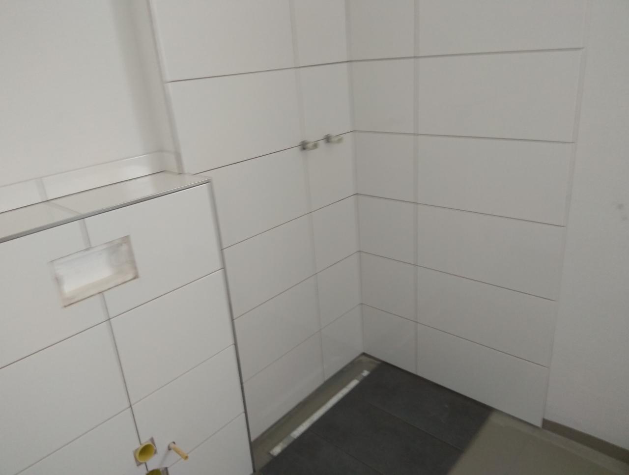 Świeżo wykafelkowana ściana łazienki z widocznymi otworami na instalacje sanitarne i fragmentem podłogi wyłożonej ciemnymi panelami.