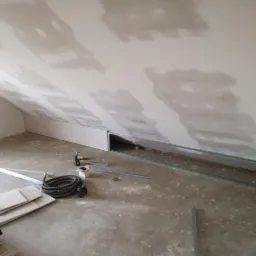 Poddasze w trakcie wykańczania: ściany i skośny sufit pokryte gładzią gipsową, widoczne profile metalowe przy podłodze, narzędzia i materiały budowlane w nieładzie.