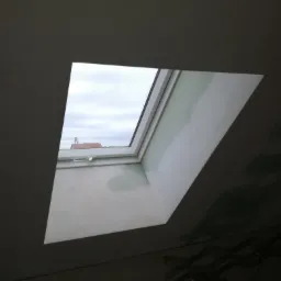 Zamontowane okno dachowe z widokiem na pochmurne niebo, widoczne ślady wilgoci na ścianie wokół okna i fragment łazienki w dolnej części kadru.