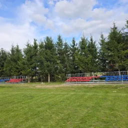 Stalowa konstrukcja trybun z czerwonymi i niebieskimi siedziskami na tle zielonego trawnika i rzędu świerków pod błękitnym niebem.