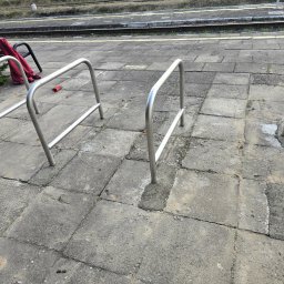 RAMIRES - Metalowe stojaki rowerowe na peronie kolejowym, przymocowane do betonowej nawierzchni, z czerwoną bluzą i rękawiczką leżącą obok ławki w tle.