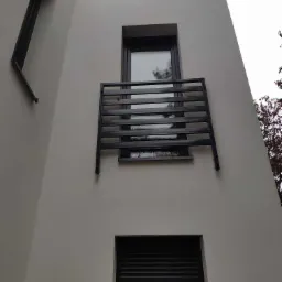 Minimalistyczna, czarna balustrada balkonowa z poziomych profili, zamontowana na elewacji budynku z widocznymi oknami i roletami.