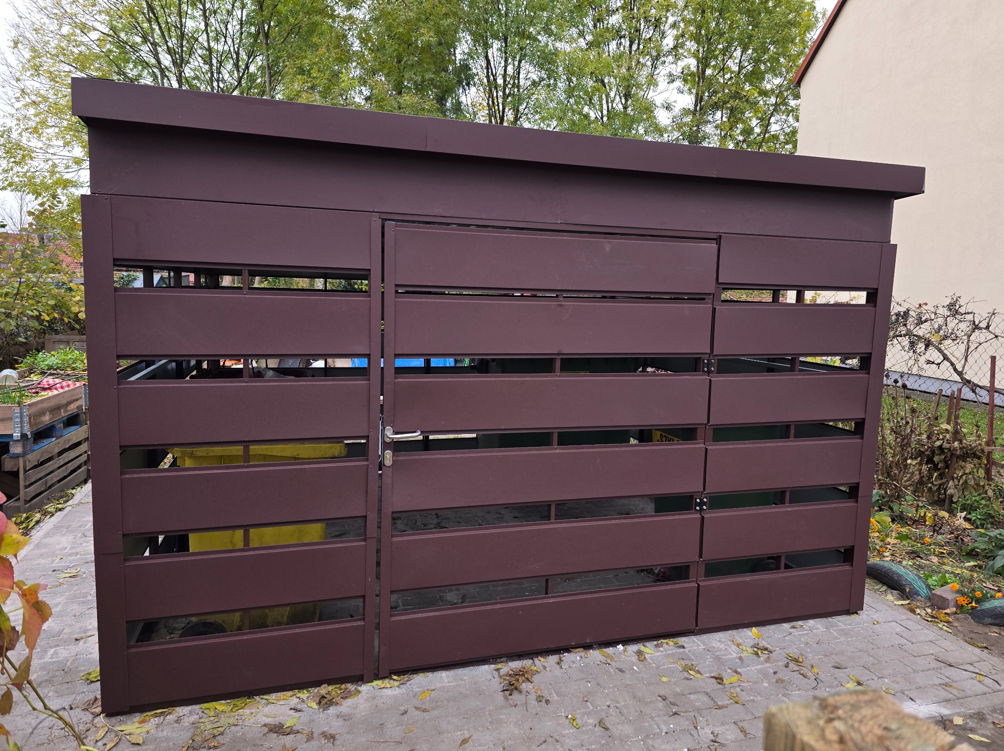 Brązowa, metalowa zabudowa śmietnikowa z poziomych listew, zamykana na klucz, na brukowanym podłożu, z widocznymi pojemnikami na śmieci wewnątrz.