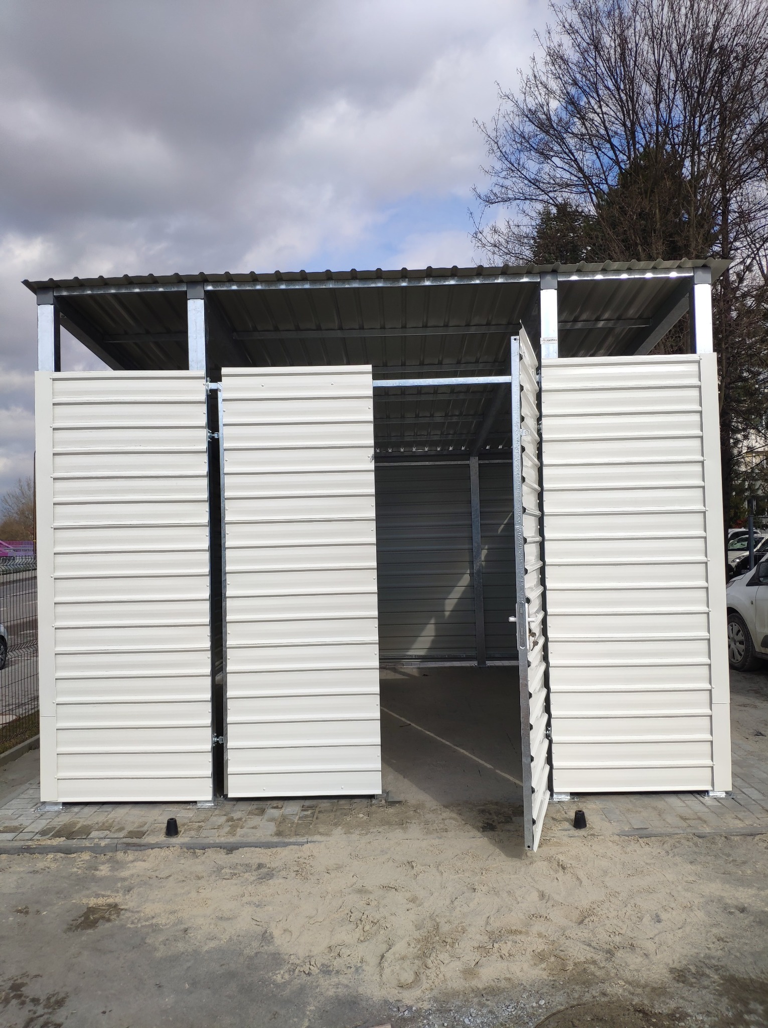 Metalowa konstrukcja z zasuwanymi, białymi panelami, prawdopodobnie na śmietniki, z otwartymi drzwiami po prawej stronie, na betonowym podłożu.