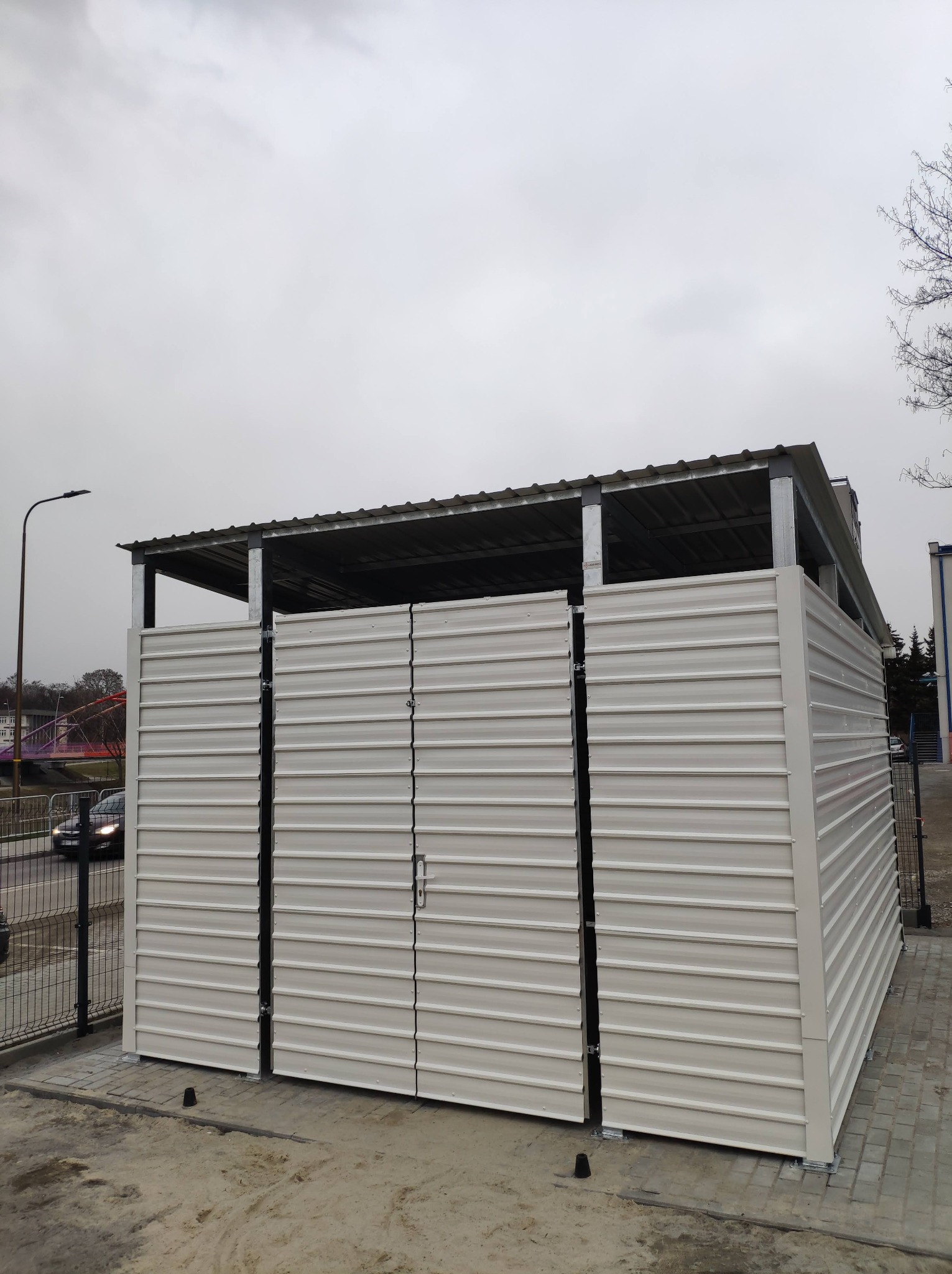 Metalowa osłona śmietnikowa z zamykanymi drzwiami, widok z zewnątrz na tle miejskiego krajobrazu z mostem i przejeżdżającymi samochodami.