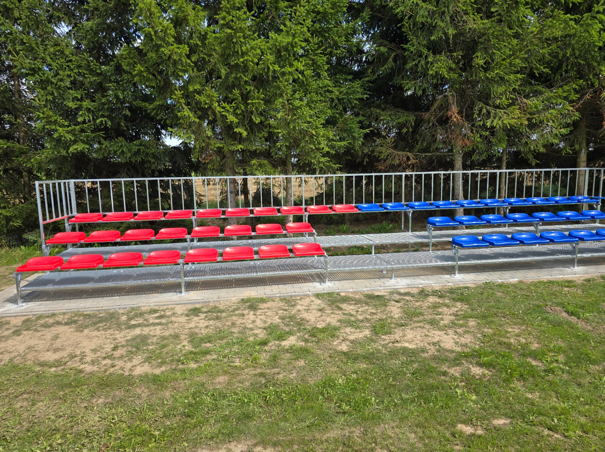Metalowa konstrukcja trybun zewnętrznych z czerwonymi i niebieskimi plastikowymi siedziskami na tle drzew iglastych.