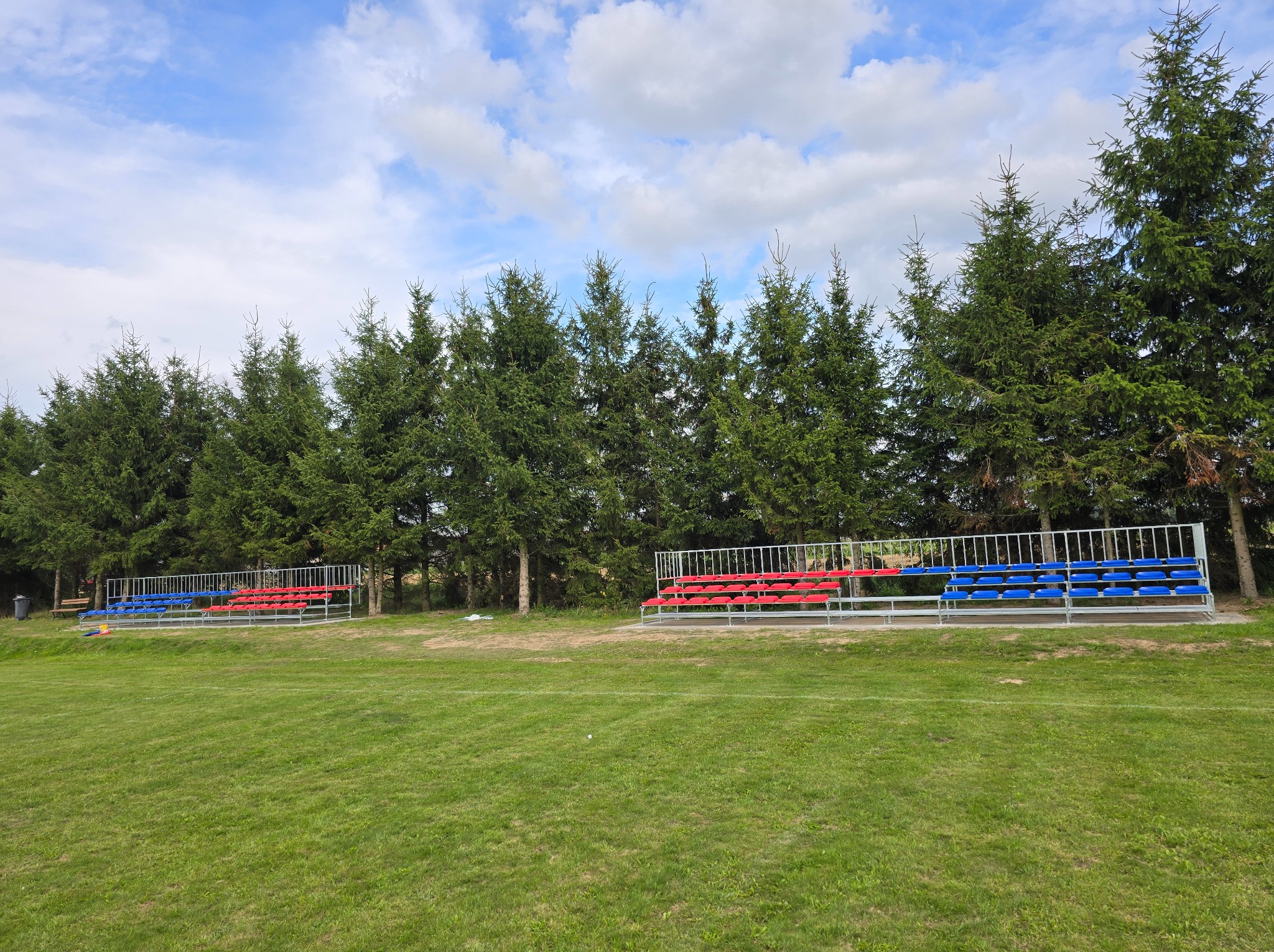 Stalowa konstrukcja trybun z czerwonymi i niebieskimi siedziskami na tle zielonego trawnika i rzędu świerków pod błękitnym niebem.