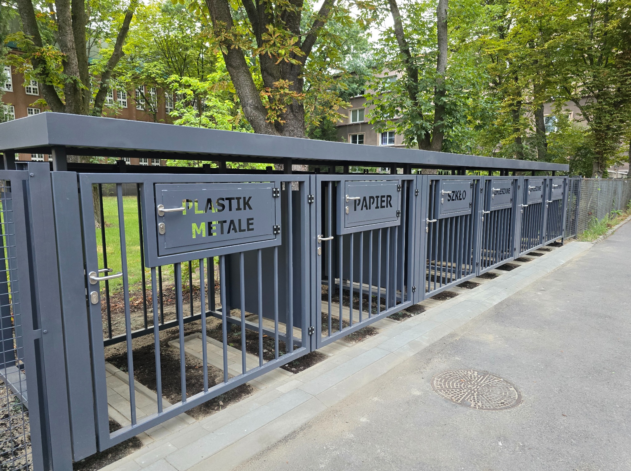 Szary metalowy system pojemników na odpady segregowane: plastik i metal, papier, szkło, zmieszane; na zewnątrz, z zielenią w tle.