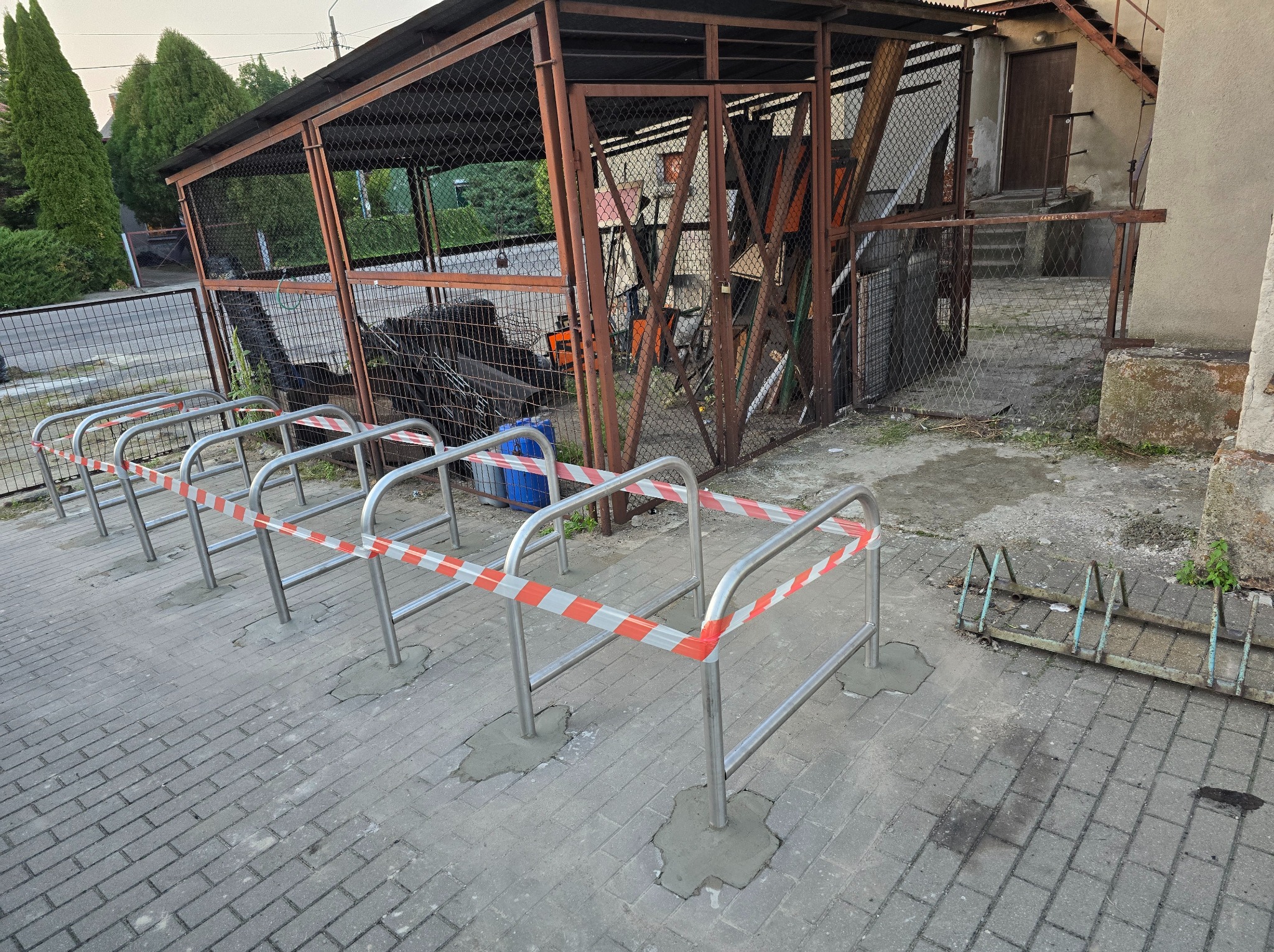 Nowa, metalowa konstrukcja stojaka na rowery, odgrodzona taśmą ostrzegawczą, obok starego, zardzewiałego stojaka, na tle metalowej klatki wypełnionej różnymi przedmiotami.