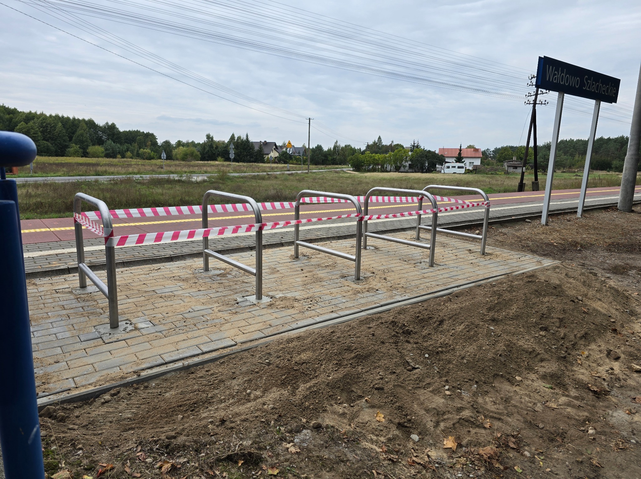 Metalowe barierki ochronne na peronie kolejowym, zabezpieczone taśmą ostrzegawczą, w tle tablica z nazwą stacji Wałdowo Szlacheckie.