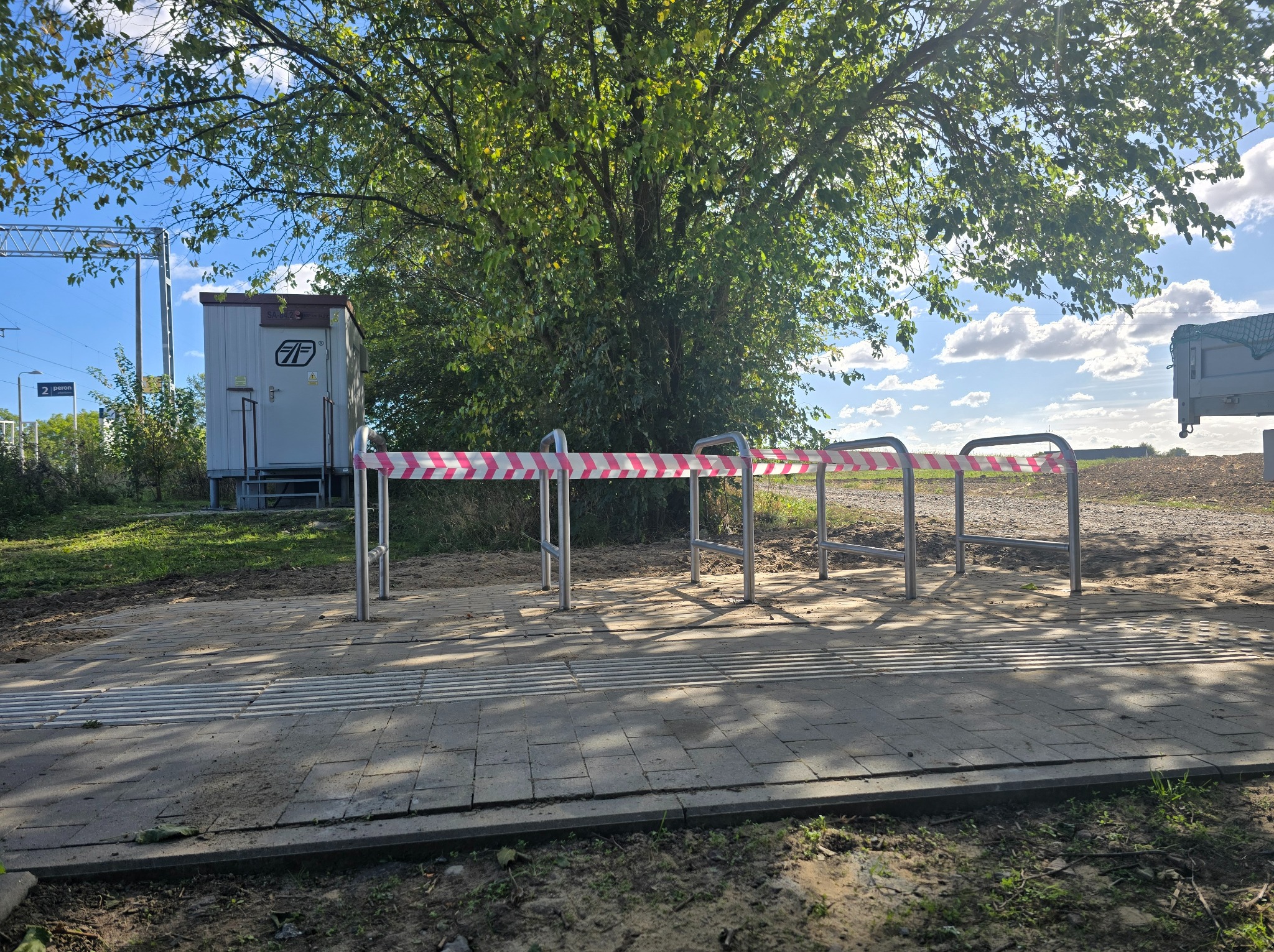 Nierdzewna poręcz zabezpieczona taśmą ostrzegawczą na tle drzewa i kontenera przy peronie.