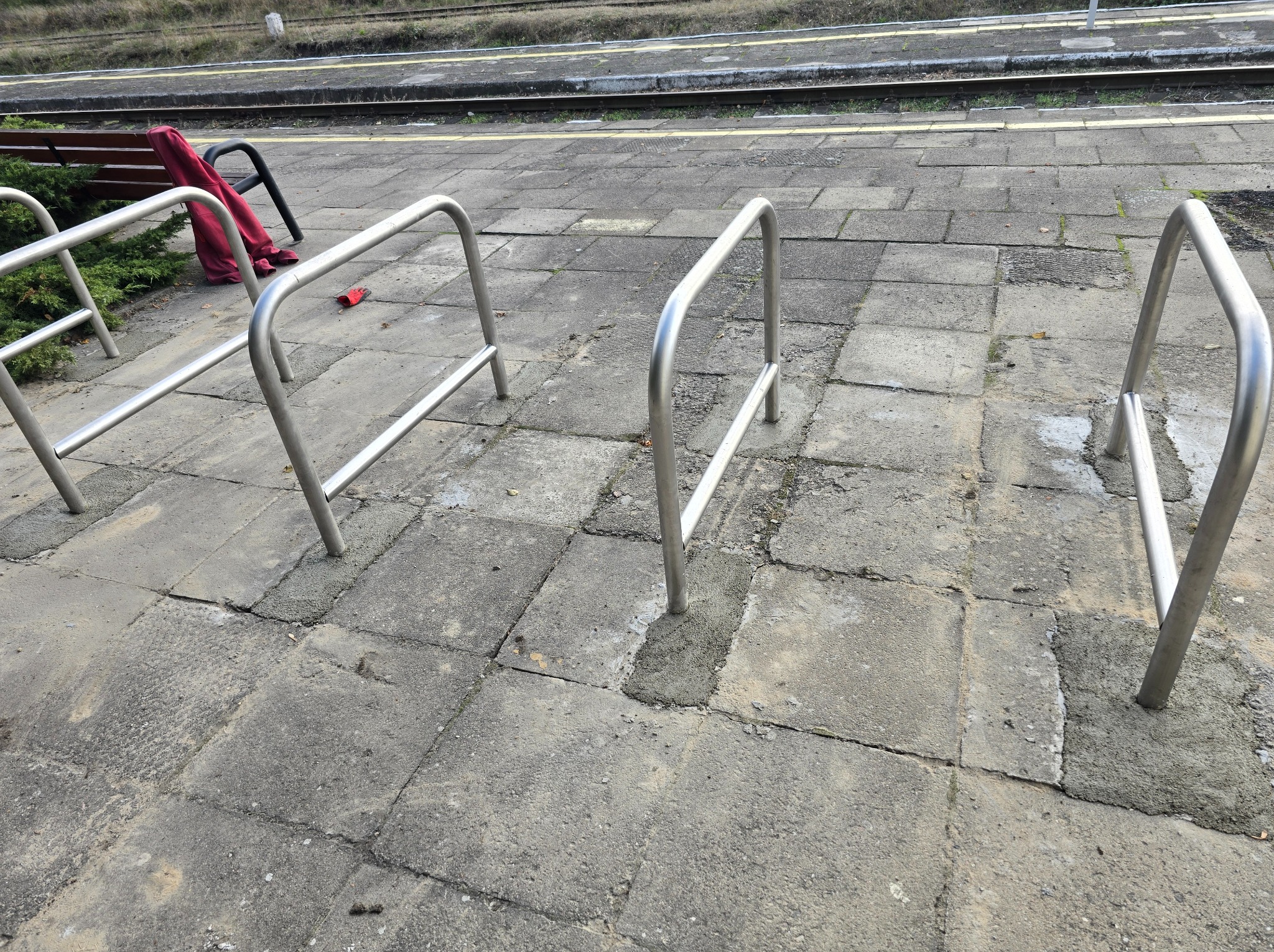 Metalowe stojaki rowerowe na peronie kolejowym, przymocowane do betonowej nawierzchni, z czerwoną bluzą i rękawiczką leżącą obok ławki w tle.