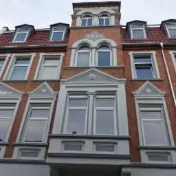 Fasada odnowionego, trzypiętrowego budynku z czerwonej cegły z szarymi detalami architektonicznymi, okna z szarymi ramami, czerwony dach z lukarnami, rok budowy 1894 widoczny na szczycie.