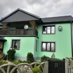 Dom jednorodzinny po remoncie elewacji, pomalowany na jasnozielony kolor, z balkonem i szarą dachówką. Widoczne anteny satelitarne na ścianie i dekoracje okienne.