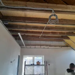 Poddasze w trakcie wykończenia: widoczne ocieplenie między krokwiami, częściowo wykończona ściana z gładzią, rusztowanie z narzędziami pod oknem dachowym i zwisające przewody elektryczne.