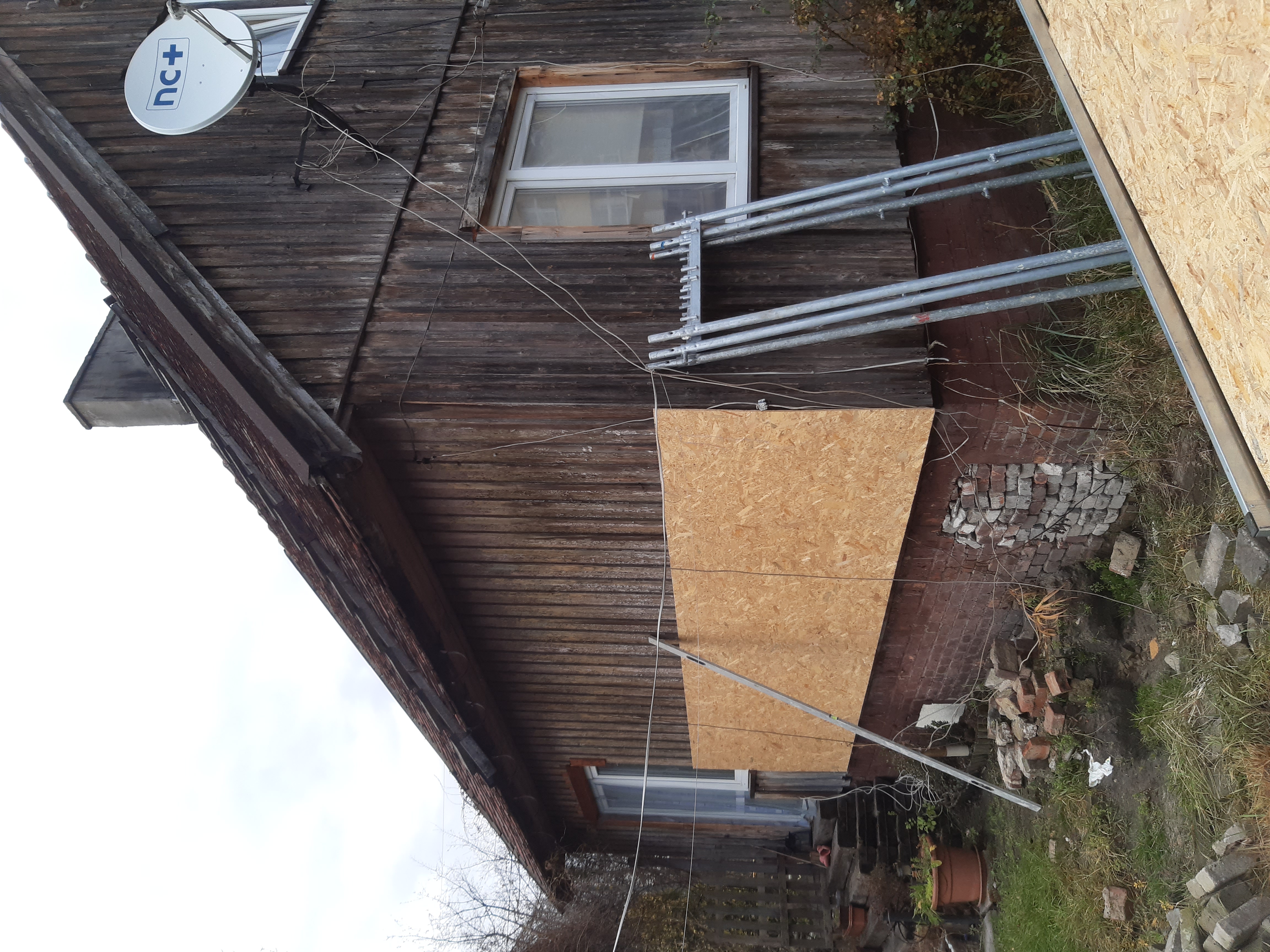 Fragment starego drewnianego domu z częściowo zamontowaną płytą OSB na elewacji, widoczne rusztowanie i sterta cegieł na ziemi.