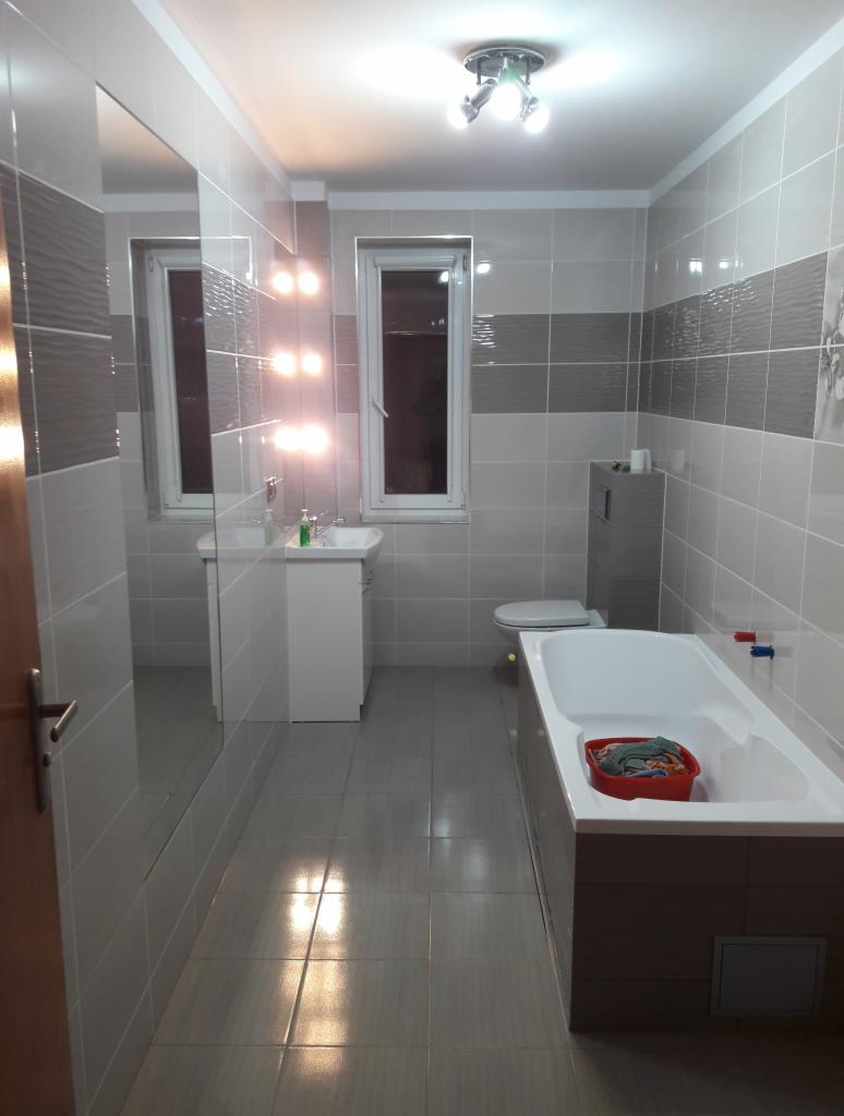 Jasna łazienka po renowacji z szarymi płytkami, białą armaturą i lustrem odbijającym okno, z praniem w wannie.