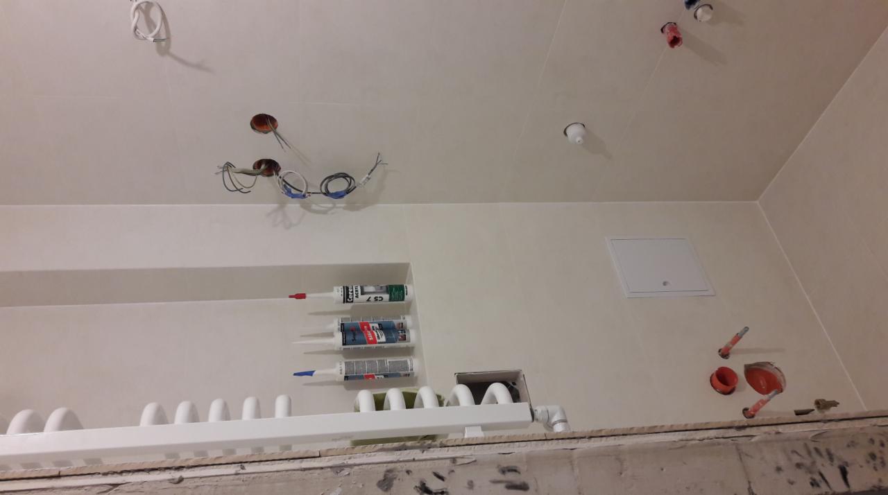 Wnętrze łazienki w trakcie prac wykończeniowych: widoczne przewody elektryczne wystające z sufitu, rury grzewcze, silikony budowlane w półce, otwory w ścianie z wystającymi rurami
