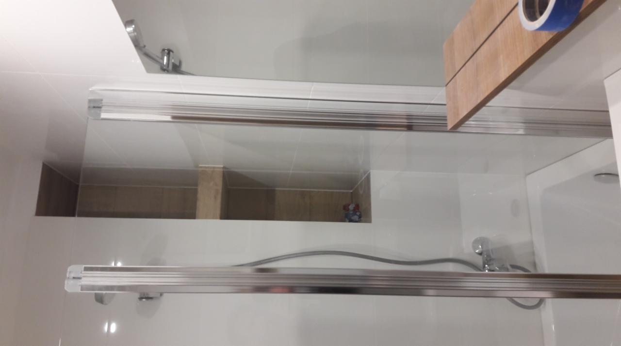 Szklana kabina prysznicowa z przesuwanymi drzwiami, widoczne białe płytki, chromowane elementy i fragment drewnianej półki z taśmą malarską.