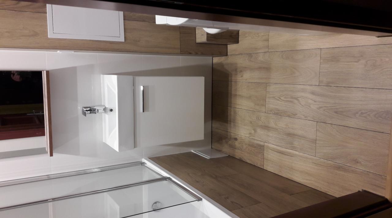 Nowoczesna łazienka z drewnopodobnymi płytkami na ścianie, białą szafką pod umywalką i kabiną prysznicową ze szklanymi drzwiami.