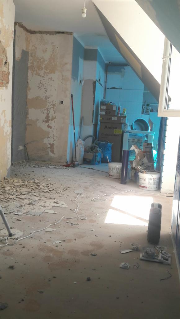 Wnętrze w trakcie remontu: ściany z fragmentami starej farby, gruz na podłodze, widoczny karton po zmywarce Amica i inne materiały budowlane.