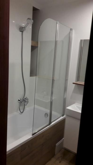 Nowoczesna łazienka z białą wanną, szklanym parawanem nawannowym, chromowaną baterią i prysznicem, oraz umywalką z szafką w minimalistycznym stylu.