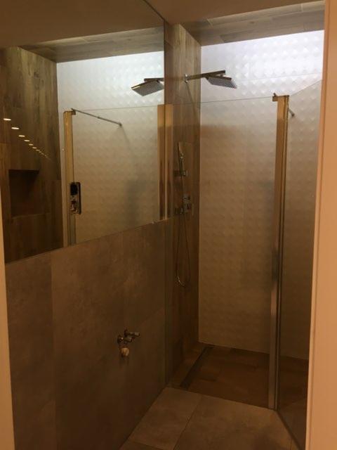 Nowoczesna kabina prysznicowa typu walk-in z dwoma deszczownicami, wykończona płytkami w odcieniach beżu i brązu, widoczna armatura i szklane ścianki.