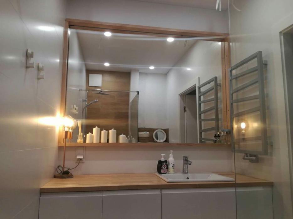 Nowoczesna łazienka z drewnianą ramą lustra, umywalką nablatową, dekoracyjnymi świecami i grzejnikiem drabinkowym, odbita w lustrze kabina prysznicowa z drewnianą ścianką.