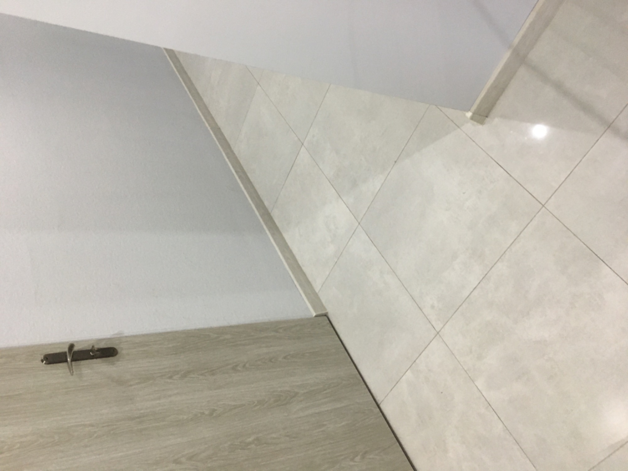 Fragment wykończonego wnętrza z jasnymi, kwadratowymi płytkami na ścianie i podłodze, białą ścianą oraz panelem imitującym drewno z zamontowanym wieszakiem na ubrania.