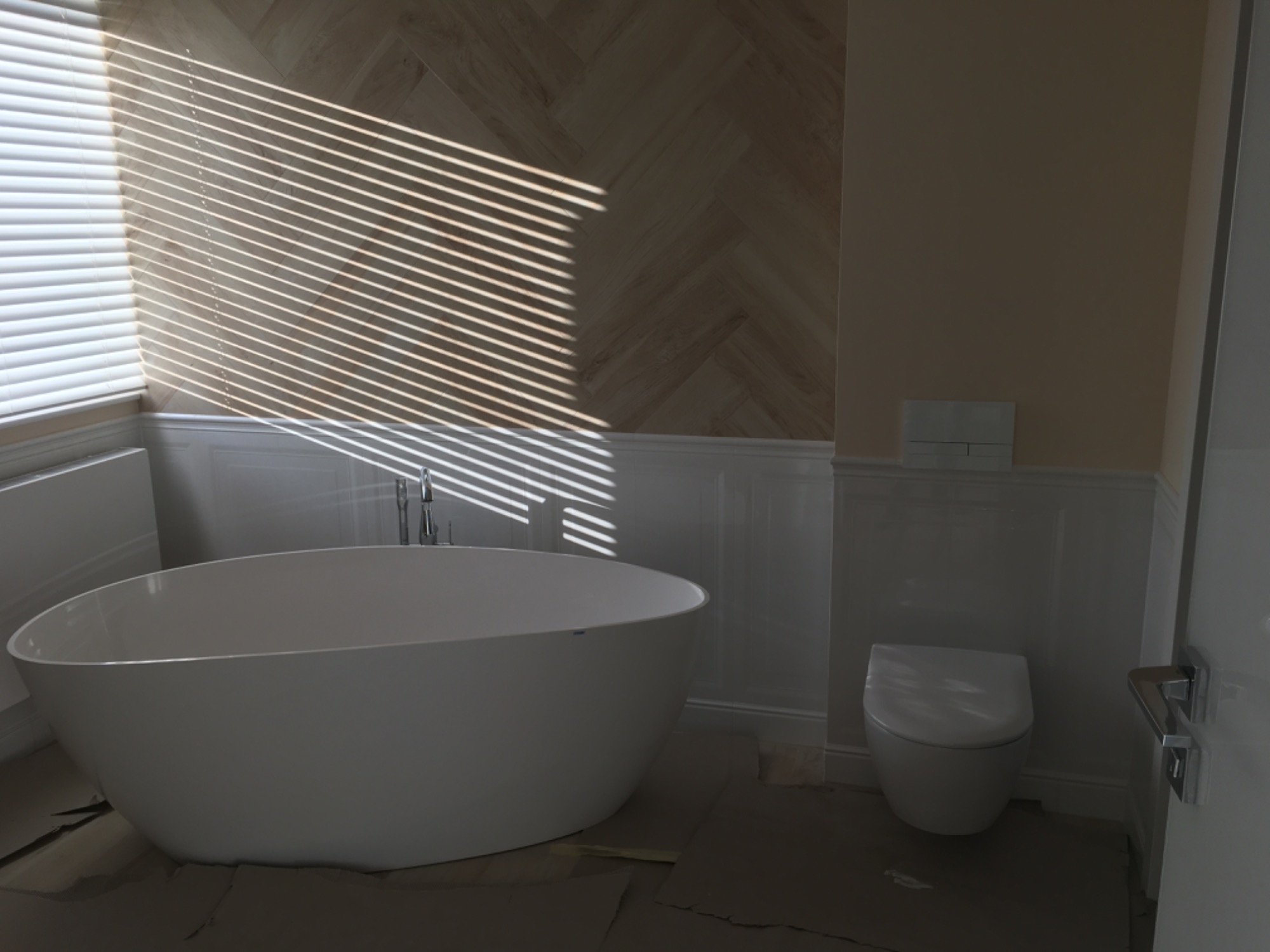 Nowoczesna, wolnostojąca wanna owalna w łazience z drewnopodobnymi płytkami ułożonymi w jodełkę, fragment toalety i promienie słoneczne wpadające przez żaluzje.