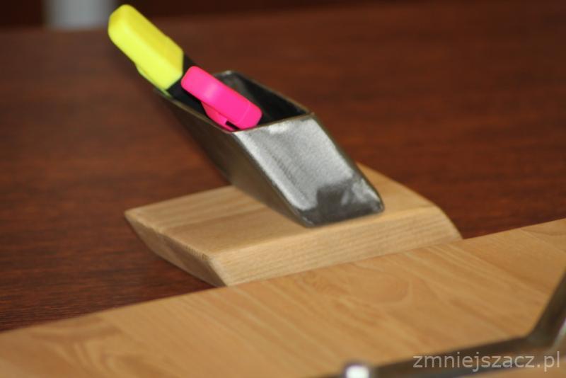 Metalowy pojemnik na drewnianej podstawie, zawierający żółty i różowy zakreślacz, ustawiony na drewnianym blacie.