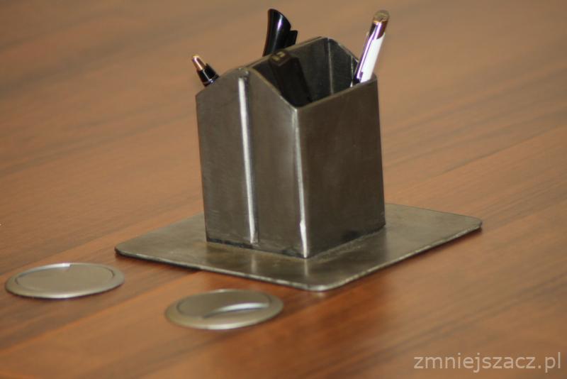 Metalowy, spawany pojemnik na długopisy, umieszczony na metalowej podstawie, stojący na drewnianym blacie biurka, obok widoczne zaślepki otworów na kable.