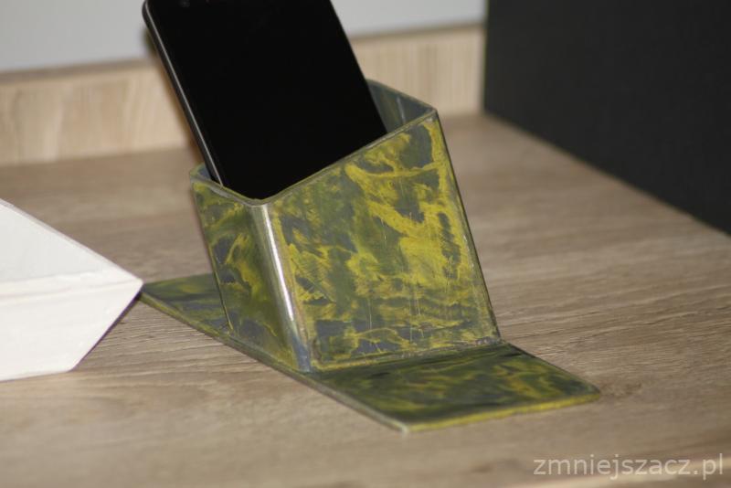 Metalowy, ręcznie wykonany stojak na telefon z żółto-zielonym, nieregularnym wzorem, umieszczony na drewnianym blacie, z czarnym telefonem w środku.