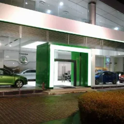 Salon samochodowy marki Skoda z widocznymi roletami zewnętrznymi nad witryną, oświetlony zielonymi neonami.