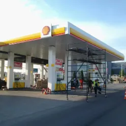 Prace na wysokości przy stacji benzynowej, widoczne rusztowanie na kółkach i pracownicy w kamizelkach odblaskowych.