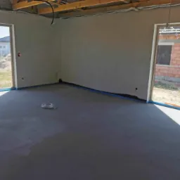 Surowe wnętrze po nałożeniu tynków na ściany i wylewki na podłogę, zabezpieczone niebieską folią okna, widoczne drewniane elementy konstrukcji dachu.